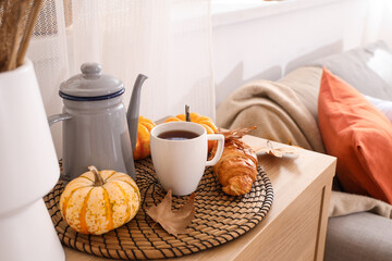 Teapot with cup of hot beverage, pumpkins, croissant and autumn leaves on table in room