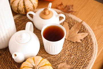 Teapot with cup of hot beverage, pumpkins and autumn leaves on table in room