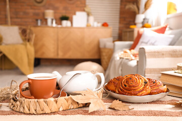 Teapot with cup of hot beverage, tasty buns and autumn leaves on table in living room