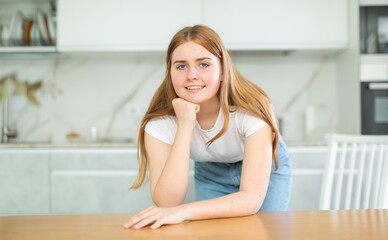 Portrait of positive teenage girl in casual clothes resting her chin on hand and leaning on countertop in cozy kitchen