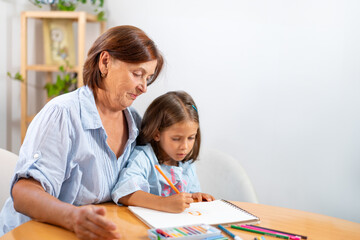 Fototapeta premium Smiling grandmother watching girl as she draws with colored pencil in book at home. The scene captures moment of admiration and encouragement as child is busy in her creative activity 