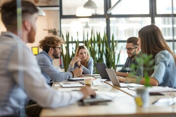 Young adults business meeting in modern office space. Caucasian colleagues sit around conference table with laptops, discuss project strategy. Female, male coworkers engage in active communication.