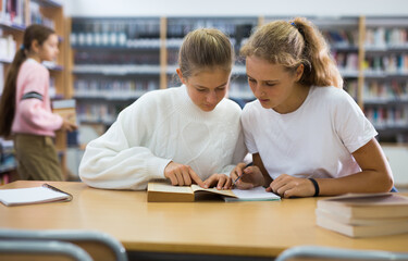 Fototapeta premium Two schoolgirls, preparing for lessons in the school library, take notes in a exercise book of the necessary material from ..the textbook, discussing topical issues