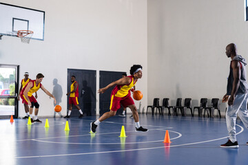 Practicing basketball drills, players dribbling around cones in gym with coach