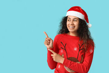 Happy young African-American woman in Christmas sweater and Santa hat pointing at something on blue background