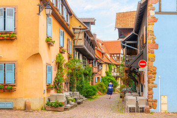 A woman in traditional dress seen from behind walking down a narrow cobblestone alley in the Alsatian medieval village of Eguisheim, France.
