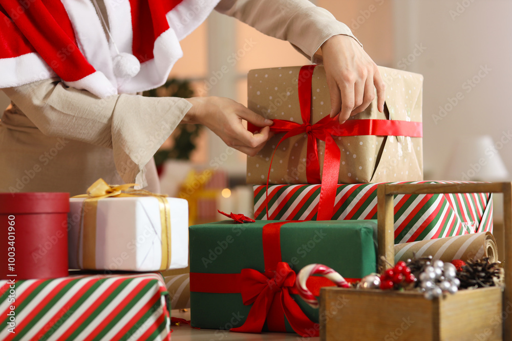 Wall mural Beautiful young woman in Santa cape tying bow while wrapping Christmas gift boxes at home, closeup