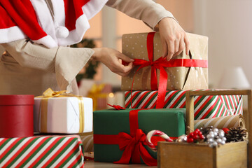 Beautiful young woman in Santa cape tying bow while wrapping Christmas gift boxes at home, closeup