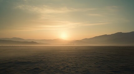 Obraz premium A hazy desert evening, with the sun setting behind a distant mountain range, casting a warm, soft light over the barren landscape.