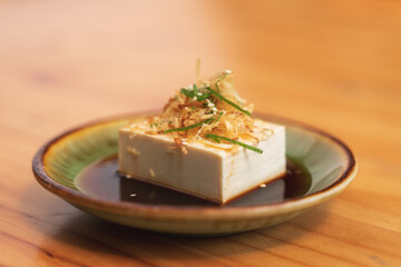 A dish of chilled tofu garnished with fried onions, sesame seeds, and green onions, served in soy sauce.