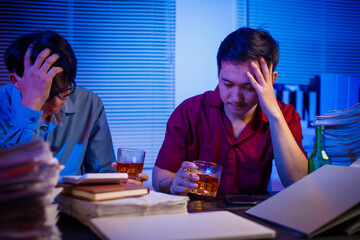 Two Asian businessmen work late into the night, sitting at a desk cluttered with a stack of papers...