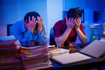 Two Asian businessmen work late into the night, sitting at a desk cluttered with a stack of papers...