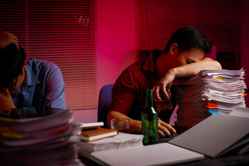 Two Asian businessmen work late into the night, sitting at a desk cluttered with a stack of papers...