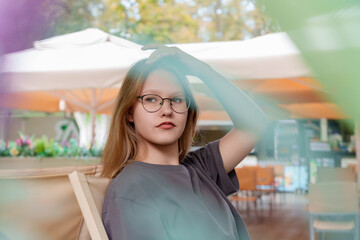 teenage girl sits alone at a summer cafe table, her attention focused inwardly as she contemplates...