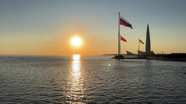 Saint-Petersburg, Russia - September 8, 2024: Horizontal 4K video. Flag of Russian Federation, USSR, Russian Empire on shores of Gulf of Finland. Setting sun. Reflection in water. Sunset. Urban view