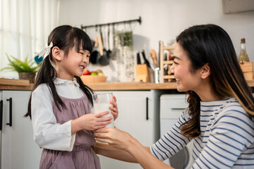 Asian little daughter drinking a glass of milk with mother in kitchen.
