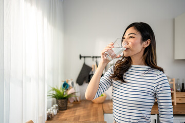 Asian young woman drinking a glass of water in kitchen at home. 