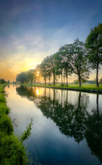 A serene sunset casts vibrant hues over a calm river, with trees reflecting in the still waters surface