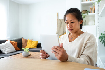 Young Asian woman working at home office holding tablet reading document with serious expression.