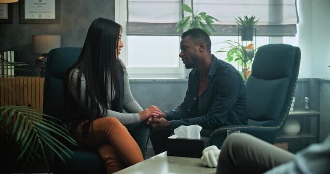 Happy multiethnic young family on counseling session with psychotherapist. Tender couple sit on chairs holding hands, laugh, find compromise after quarrel on couples therapy. Psychology concept.