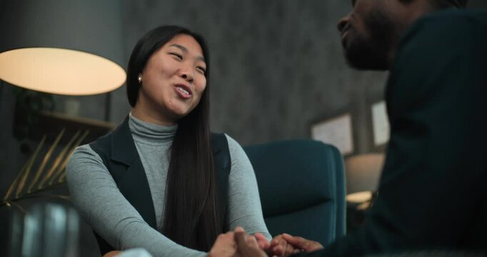 Asian woman smiles, talks to her boyfriend or husband during counseling session with therapist. Happy multiethnic couple sit on chairs holding hands, find compromise after quarrel on couples therapy.