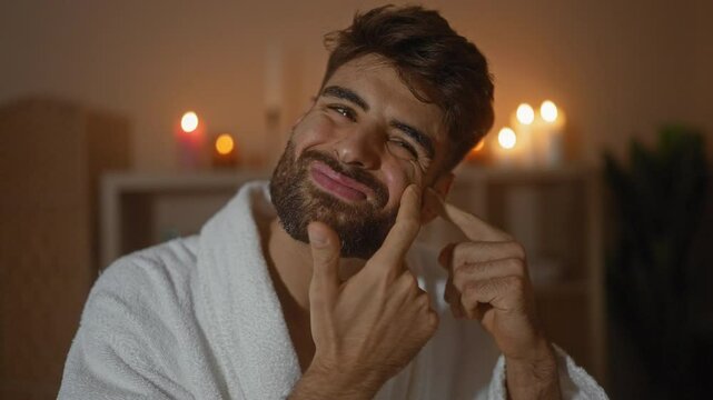 Handsome man with beard in spa room wearing white robe and squeezing a pimple on his face with candles glowing in the background for a wellness treatment