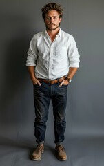 A man in a white shirt and blue jeans stands in front of a gray background. He is wearing a brown belt and a watch