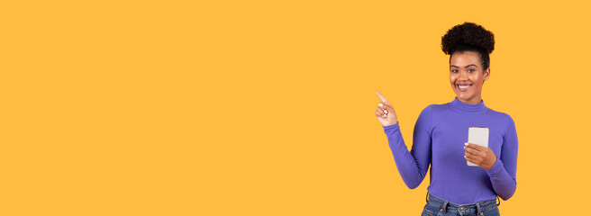 A joyful woman in a purple top stands against a vibrant yellow backdrop, holding a smartphone in one hand while playfully pointing at copy space, creating a cheerful and engaging atmosphere.