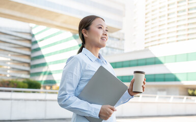 Asian woman stands outdoors, smiling while holding a laptop and a coffee cup. She is dressed in a collared shirt and appears relaxed in a modern urban setting on a clear day.