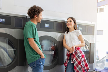A young man and woman share a light-hearted conversation at a laundromat, surrounded by washing machines. The atmosphere is relaxed and friendly as they wait for laundry.