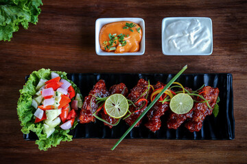 Fried Pork pieces in a black bowl along with lemon slices placed in a dark wooden backdrop separately placed with mayonnaise, mint flavored sauces and vegetables salad