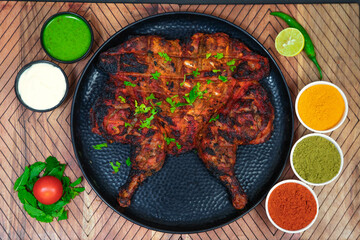 Tandoori chicken, full barbeque chicken along with mayonnaise, onion slice, mint leaves, chili powder, coriander powder, turmeric powder and spices on black plate in the wooden background