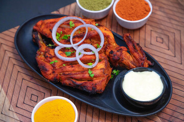 Tandoori chicken, barbeque chicken along with mayonnaise, onion slice, mint leaves, chili powder, coriander powder, turmeric powder and spices on black plate and black bowl in the wooden background