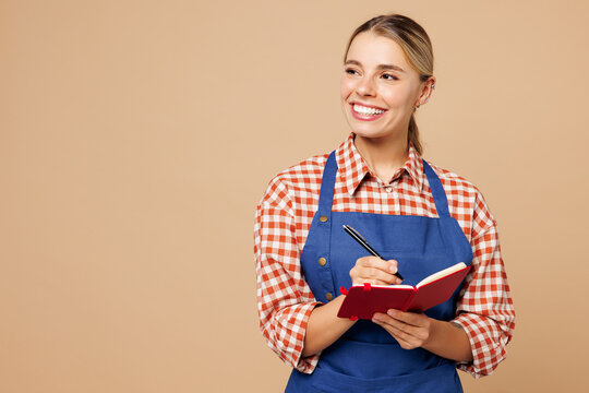 Young woman barista bartender barman employee wear blue apron red shirt work in coffee shop write down order in notebook look aside isolated on plain beige background. Small business startup concept.