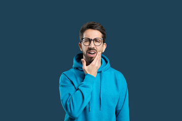 A man wearing a blue hoodie displays a pained expression while holding his chin. The dark background adds focus to his discomfort, possibly indicating a toothache or jaw pain.