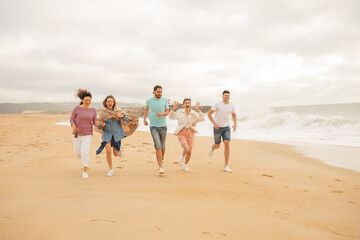 A group of five friends run joyfully along the beach, laughing and embracing the warm atmosphere as waves crash nearby during a cloudy sunset.