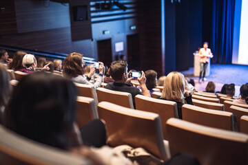 Business and entrepreneurship symposium. Female speaker giving a talk at business meeting. Audience in conference hall. Rear view of unrecognized participant in audience.