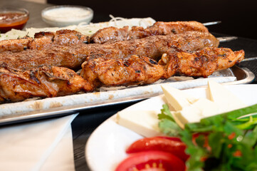 Close-up of grilled meat skewers served on a platter with fresh vegetables and cheese on the side, accompanied by sauces