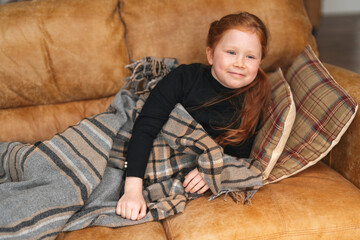 Fototapeta premium Cozy moments on a chilly afternoon with a young girl wrapped in a blanket on a comfortable couch at home