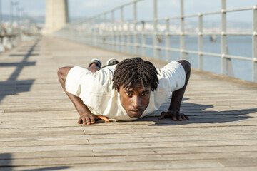 African American young man is performing push-ups on a wooden pier overlooking the calm water. The sun shines brightly, highlighting his focused expression and dedication to fitness.
