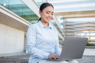Fototapeta premium A young Asian woman smiles while working on her laptop outside a contemporary office building. She is focused and appears to enjoy her task in the pleasant environment.