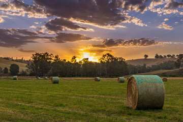 The sun sets over a tranquil rural landscape, casting warm hues across the sky while hay bales are arranged in a lush green field, creating a picturesque scene.