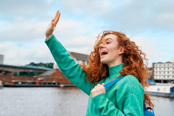 A joyful young woman waves enthusiastically by the riverside, surrounded by a picturesque cityscape...