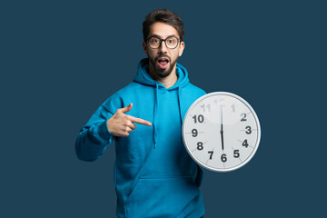 A young man wearing a blue hoodie stands against a solid background, expressing surprise while...