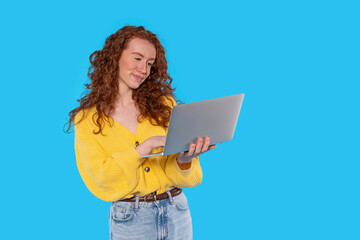 A young woman with curly hair smiles while working on her laptop against a vibrant blue background in a cozy setting