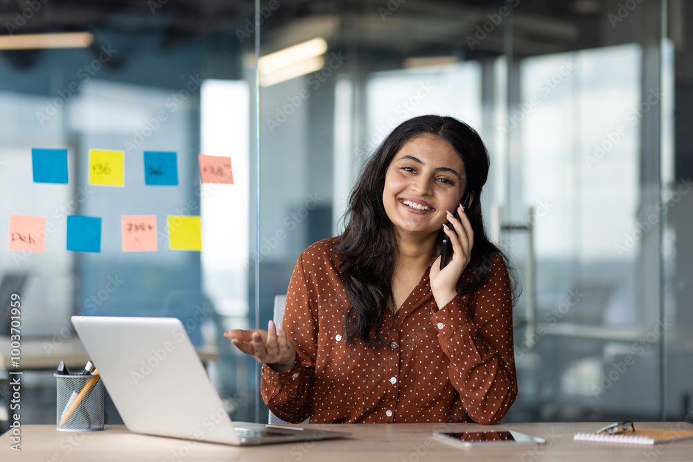 Poster Woman smiling talking phone in modern office. Laptop on desk with colorful sticky notes on glass wall, conveying productivity and organization. Bright atmosphere highlighting professional environment