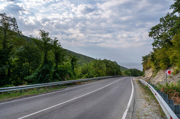 Road landscape forest, mountains and sea on the horizon. Empty long mountain road to the horizon on a sunny summer day with dramatic cloudy sky. Highway turn panoramic view.