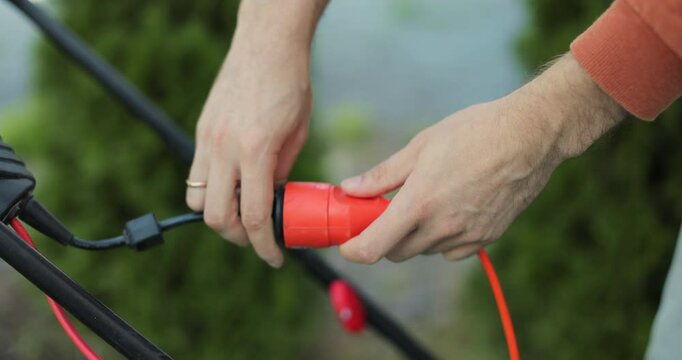 Man plugs in an electric lawnmower. Close-up of a man hands plugging the cable into the outlet.