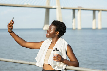 A cheerful young African American woman enjoys a moment by the water, capturing a selfie with a smartphone while holding a water bottle. The bridge in the background adds to the scenic view.