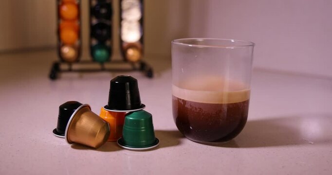 A clear glass filled with freshly brewed coffee appears alongside coffee capsules, showcasing a neat capsule organizer in the background
